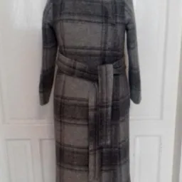 Szary, wełniany płaszcz w kratę z paskiem, prezentowany na manekinie na tle białych drzwi.