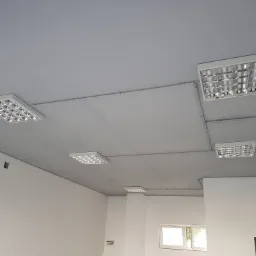 Sufit w szarym odcieniu z zamontowanymi kwadratowymi lampami rastrowymi połączonymi rurkami instalacyjnymi; w tle widoczne białe ściany i drabina aluminiowa.