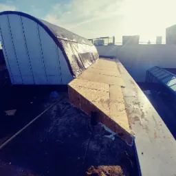 Konstrukcja dachu z elementami drewnianymi i pokryciem z folii, widok z góry na tle słonecznego nieba i zabudowań miejskich.