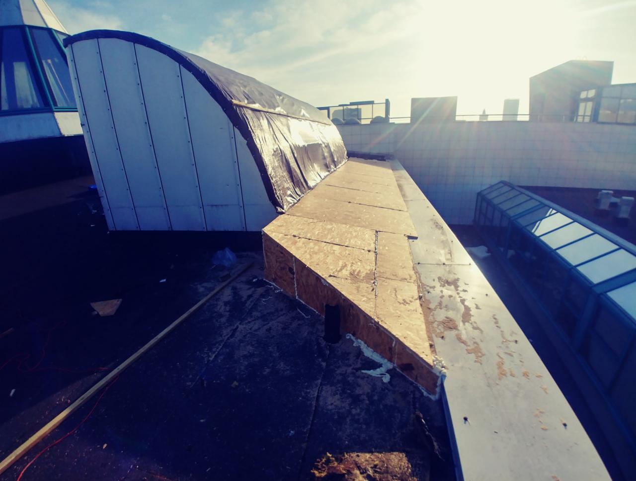 Konstrukcja dachu z elementami drewnianymi i pokryciem z folii, widok z góry na tle słonecznego nieba i zabudowań miejskich.