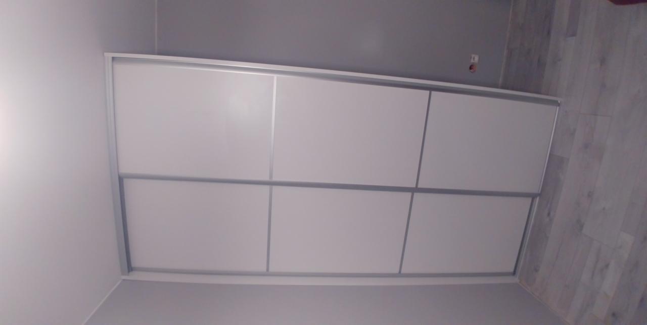 Biała szafa przesuwna z sześcioma panelami, wbudowana w szarą ścianę, widoczna pod kątem, z fragmentem drewnopodobnej podłogi.