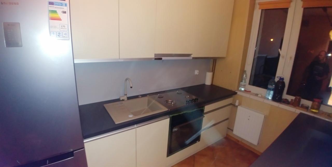 Jasna zabudowa kuchenna z ciemnym blatem, widoczny zlew, płyta gazowa i piekarnik, obok lodówka Samsung z etykietą energetyczną, okno z widokiem na nocne otoczenie.