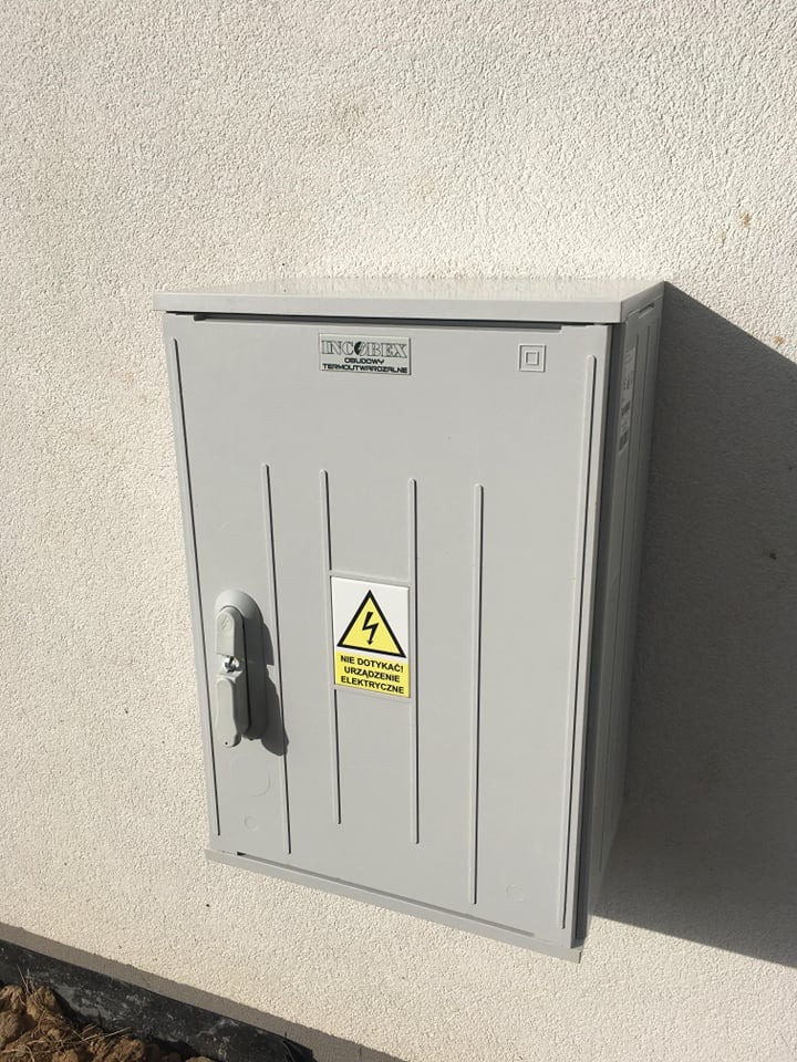 Szara skrzynka elektryczna zamontowana na białej, chropowatej ścianie budynku, z widocznym ostrzeżeniem o wysokim napięciu i napisem 'Nie dotykać urządzenia elektryczne'.