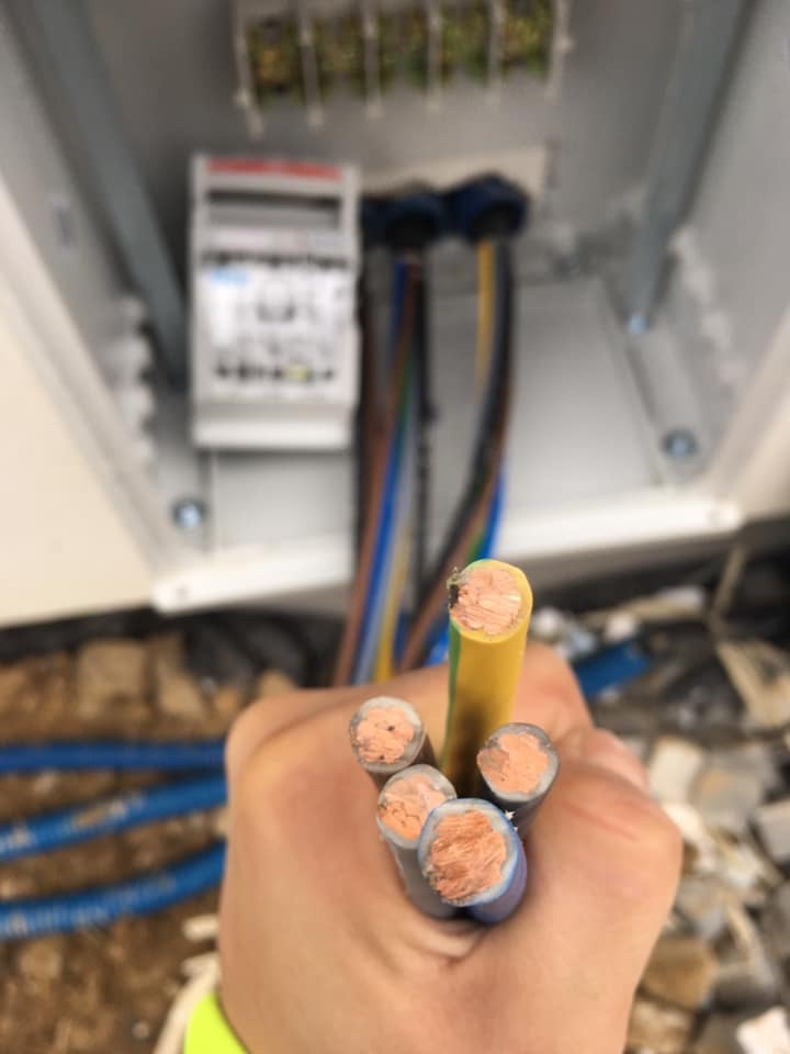 Przygotowane do podłączenia przewody elektryczne w dłoni, z widoczną skrzynką rozdzielczą w tle.