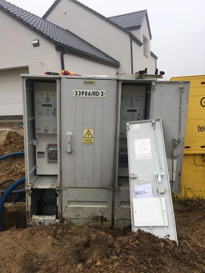 Otwarta skrzynka elektryczna z licznikami na zewnątrz budynku w trakcie podłączania, widoczny kabel w ziemi oraz narzędzia na obudowie.
