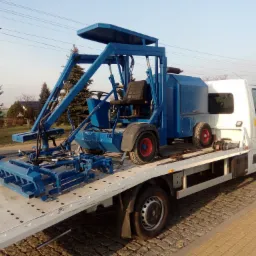 Niebieska maszyna do prac drogowych z czerwonymi felgami, umieszczona na platformie białego samochodu dostawczego z żółtymi napisami 'Pomoc Drogowa', zaparkowanego na kostce brukowej.