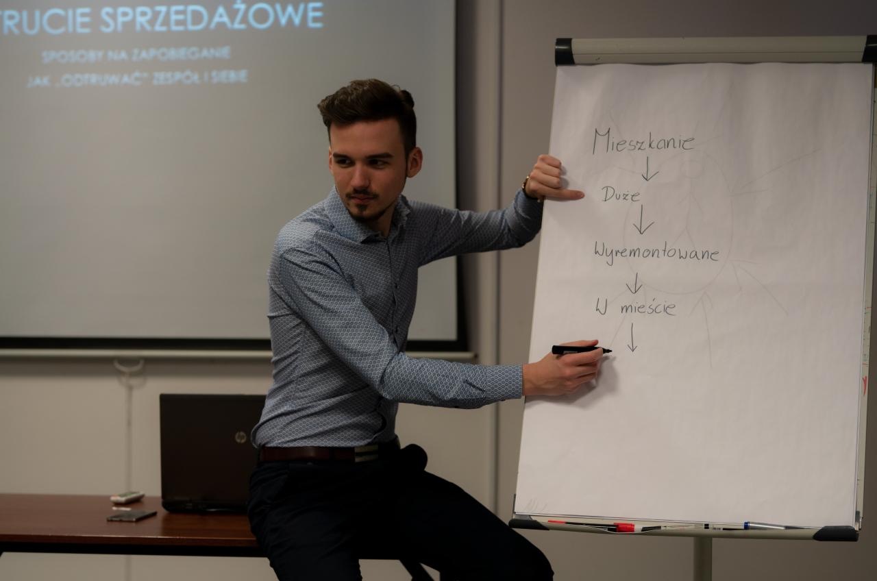 Prezenter w koszuli w geometryczny wzór, siedząc na krawędzi stołu, pisze markerem na flipcharcie z napisem 'Mieszkanie -> Duże -> Wyremontowane -> W mieście', w tle ekran projekcyjny z częściowo...