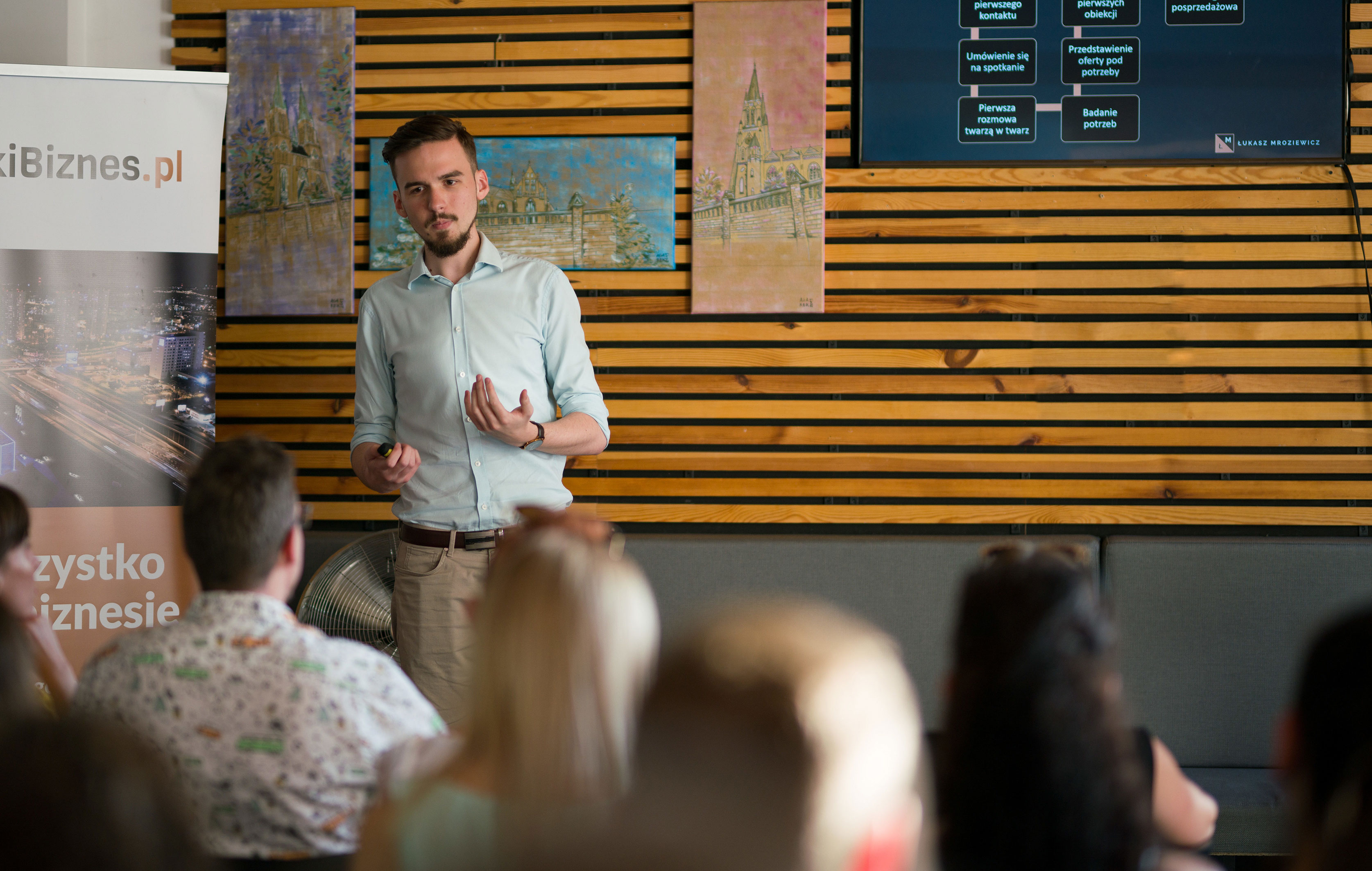 Prelegent z brodą gestykuluje podczas prezentacji przed grupą słuchaczy, w tle drewniana ściana z obrazami i ekran z hasłami sprzedażowymi.