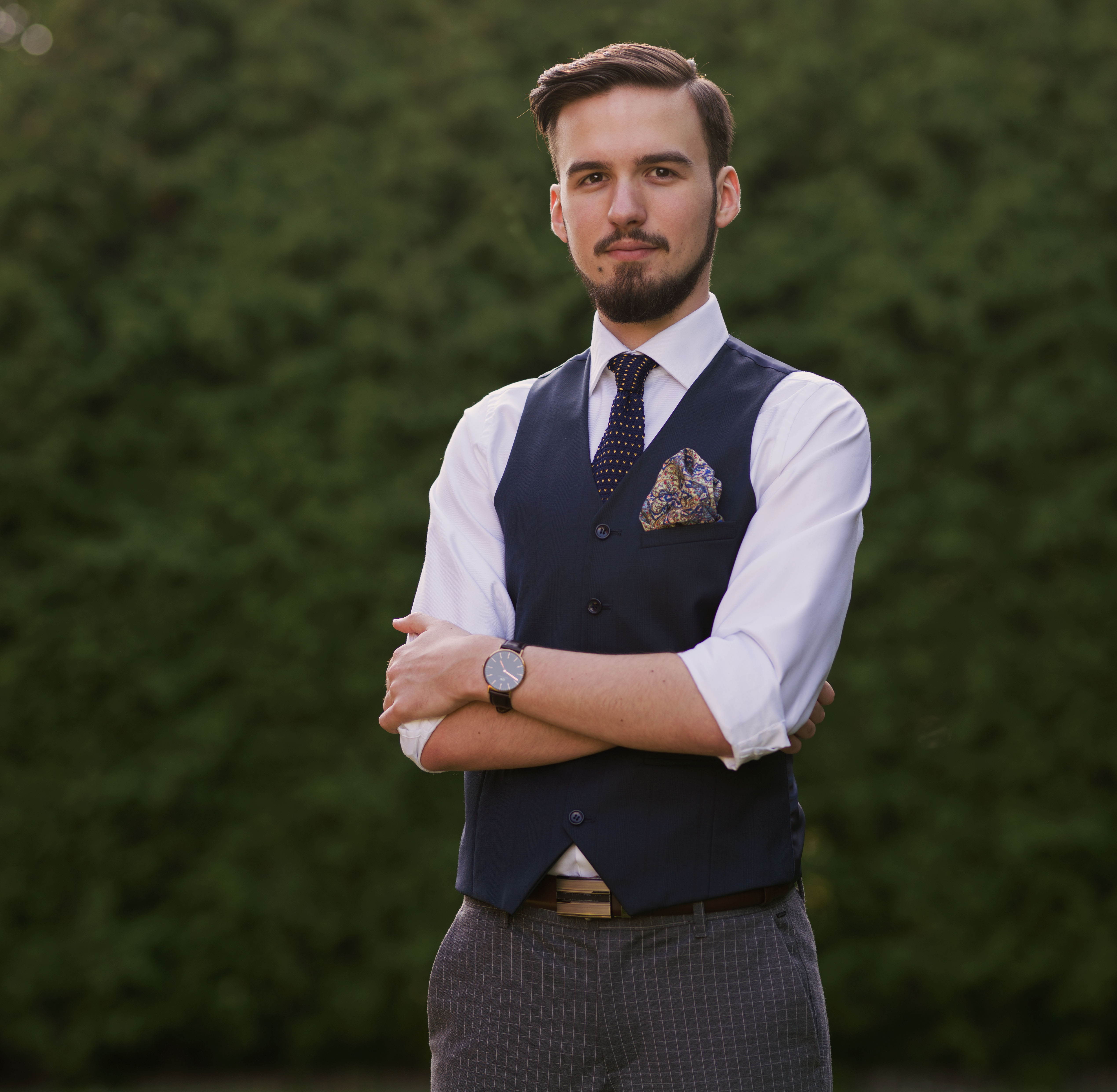 Młody mężczyzna z brodą, ubrany w białą koszulę, granatową kamizelkę, wzorzysty krawat i poszetkę, pozuje z założonymi rękami na tle zielonego żywopłotu.