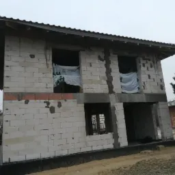 Surowy stan domu murowanego z białych bloczków w Toruniu, z widocznymi otworami okiennymi zabezpieczonymi folią, na etapie budowy.