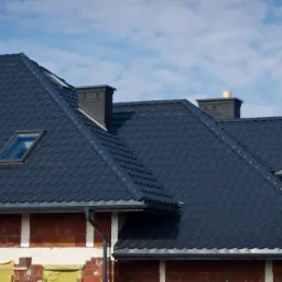 Ciemna dachówka na nowym dachu domu w trakcie budowy, widoczne rynny i okno dachowe, na dachu dekarz. Niebieskie niebo w tle.