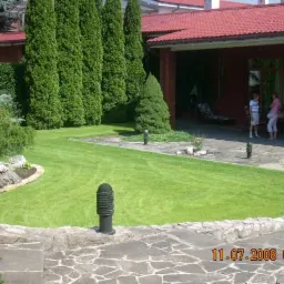 Zadbany trawnik z nasadzeniami drzew i krzewów ozdobnych, widok na taras domu z dwoma osobami w tle.