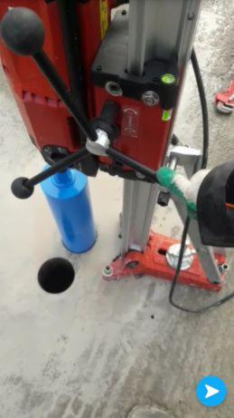 Wykonanie otworu w betonowej posadzce za pomocą wiertnicy rdzeniowej z chłodzeniem wodnym, widoczny otwór i niebieski zbiornik na wodę.