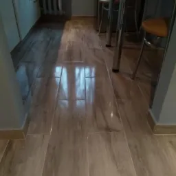 Ułożone, błyszczące panele podłogowe imitujące drewno w kuchni, widoczne fragmenty mebli i krzesła barowe