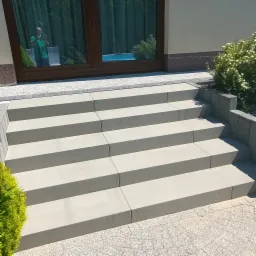 Nowoczesne schody zewnętrzne wyłożone jasnoszarą kostką brukową, z widocznym odbiciem osoby robiącej zdjęcie w przeszklonych drzwiach wejściowych, otoczone roślinnością.