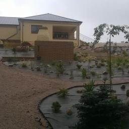 Bruk-Gard Project - Nowo założony ogród z rozłożoną agrowłókniną, nasadzeniami roślin, kamieniami dekoracyjnymi i ścieżką z kruszywa, na tle domu z ceglanym grillem.