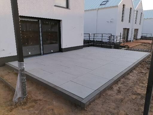 Nowoczesny taras wyłożony jasnoszarą kostką brukową w geometryczny wzór, obrzeża z ciemniejszej kostki, widok na biały budynek z dużymi oknami przesuwnymi i sąsiednie domy z metalowymi balustradami.