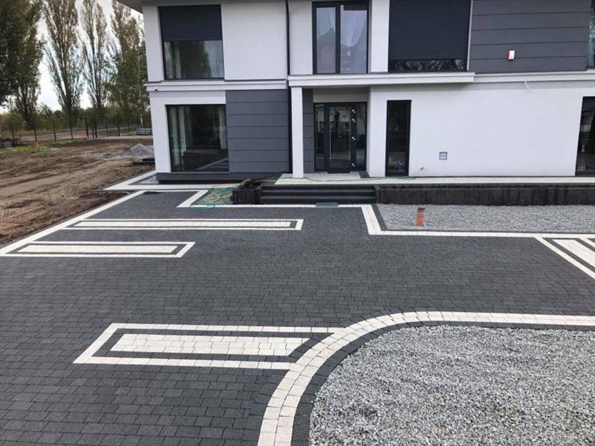 Nowoczesny podjazd z ciemnej kostki brukowej, ozdobiony białymi obramowaniami, prowadzący do dwukondygnacyjnego domu z szarą elewacją. Fragment żwirowego rabatu przy domu.