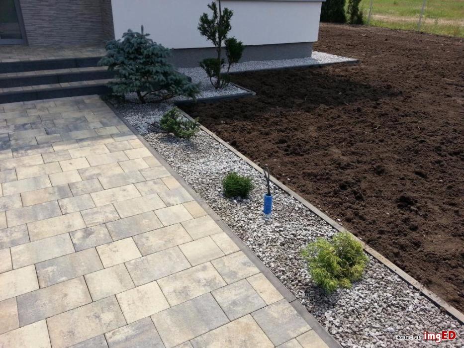 Ułożona kostka brukowa prowadząca do wejścia domu, obok rabata z kamykami i młodymi drzewkami iglastymi, świeżo przekopana ziemia w tle.