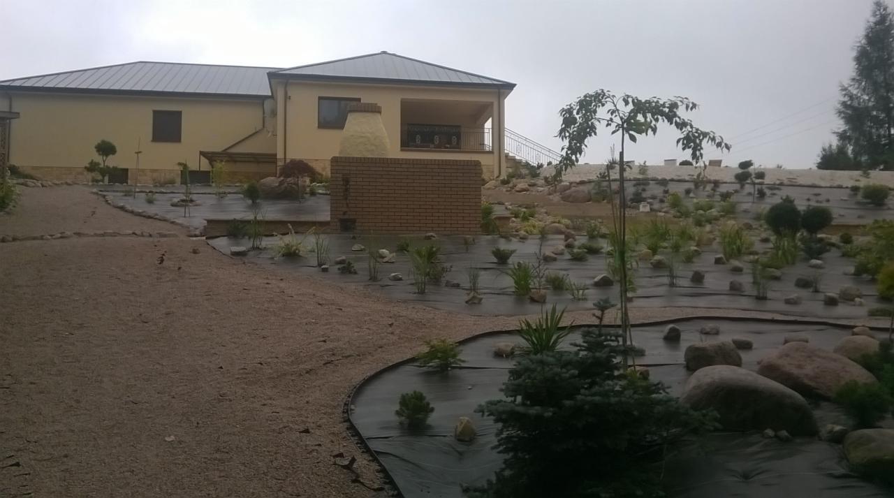 Nowo założony ogród z rozłożoną agrowłókniną, nasadzeniami roślin, kamieniami dekoracyjnymi i ścieżką z kruszywa, na tle domu z ceglanym grillem.