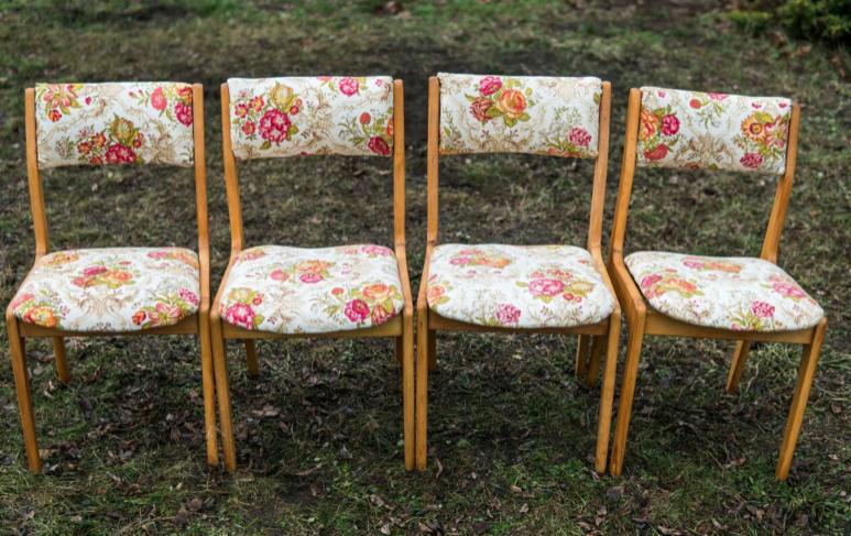 Cztery odnowione krzesła z jasnego drewna z tapicerką w kwiaty, ustawione w rzędzie na trawie.
