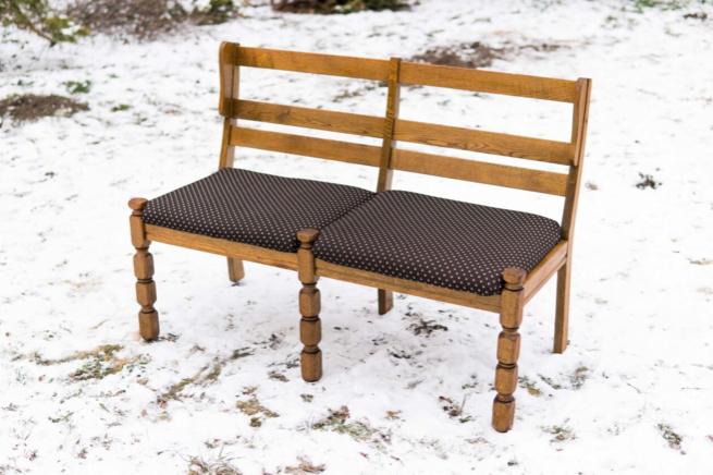 Odnowiona drewniana ławka dwuosobowa z tapicerowanymi siedziskami w brązowe kropki, ustawiona na śniegu, z widocznymi detalami toczonych nóg.