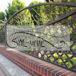 Fragment ozdobnego, metalowego ogrodzenia z motywem kwiatowym, zamontowanego na niskim murku z czerwonej cegły. W tle widoczne drzewa.