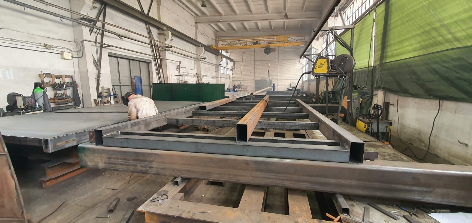 Spawanie metalowej konstrukcji szkieletowej w hali produkcyjnej, widoczny spawacz w kombinezonie i masce, suwnica pod stropem, narzędzia i materiały w tle.