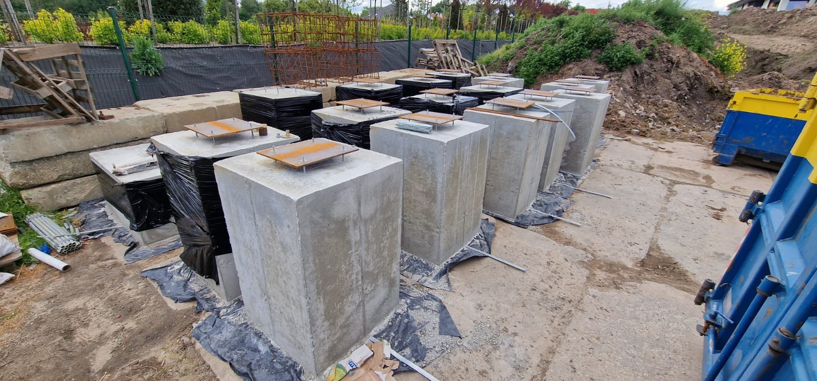 Przygotowane betonowe fundamenty pod budowę, z zamontowanymi metalowymi płytami mocującymi, częściowo zabezpieczone folią, widoczne materiały budowlane i kontener.