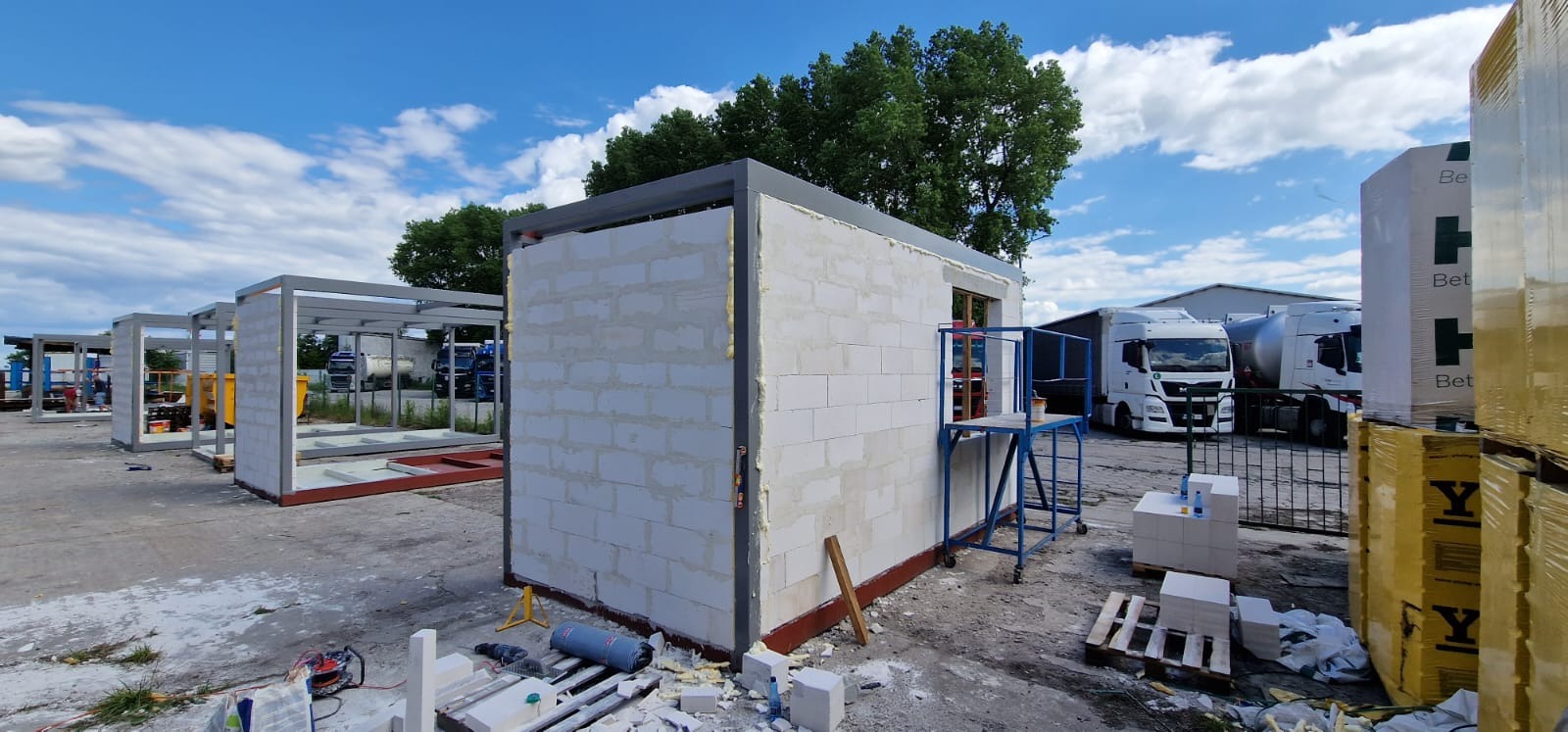 Wznoszenie ścian z bloczków betonu komórkowego na stalowej konstrukcji modułowej; widoczna drabina, palety z materiałami budowlanymi i ciężarówki w tle.