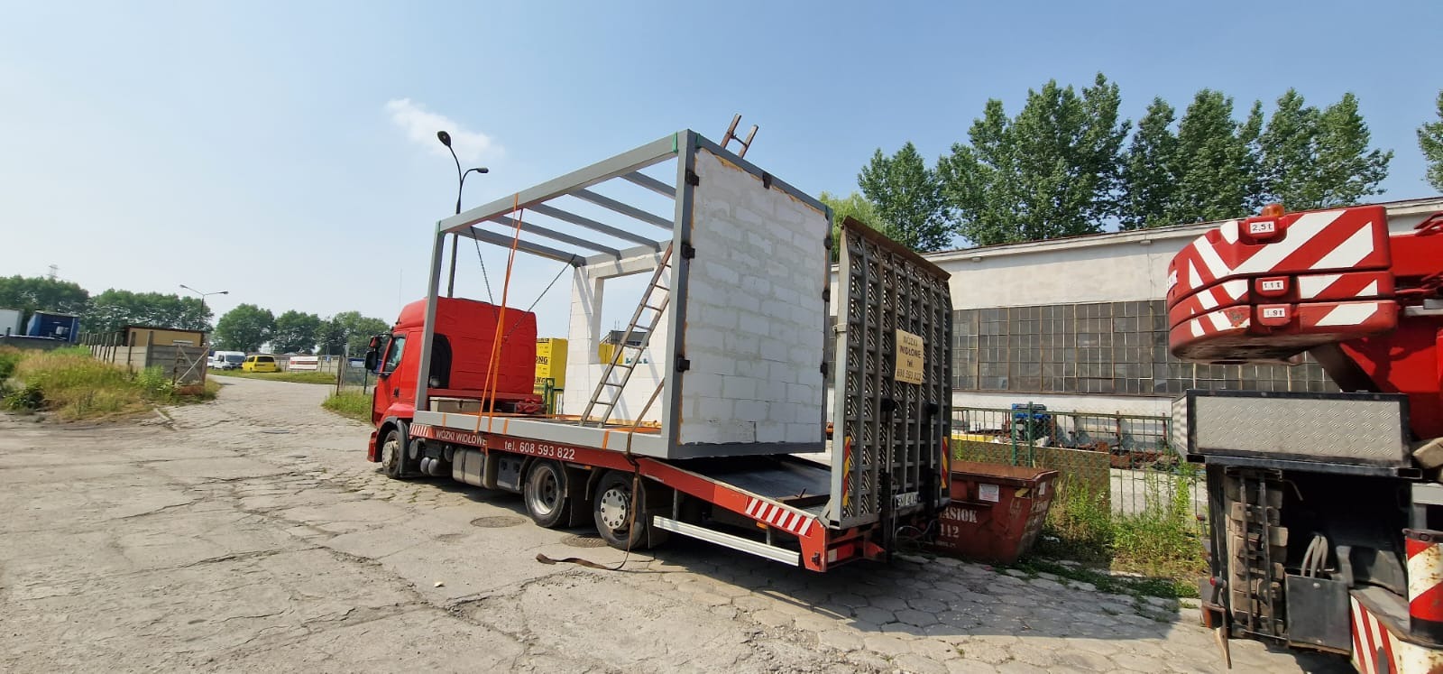 Transport modułowego domu w stanie surowym otwartym na lawecie czerwonego samochodu ciężarowego, widoczna drabina oparta o ścianę, teren przemysłowy.