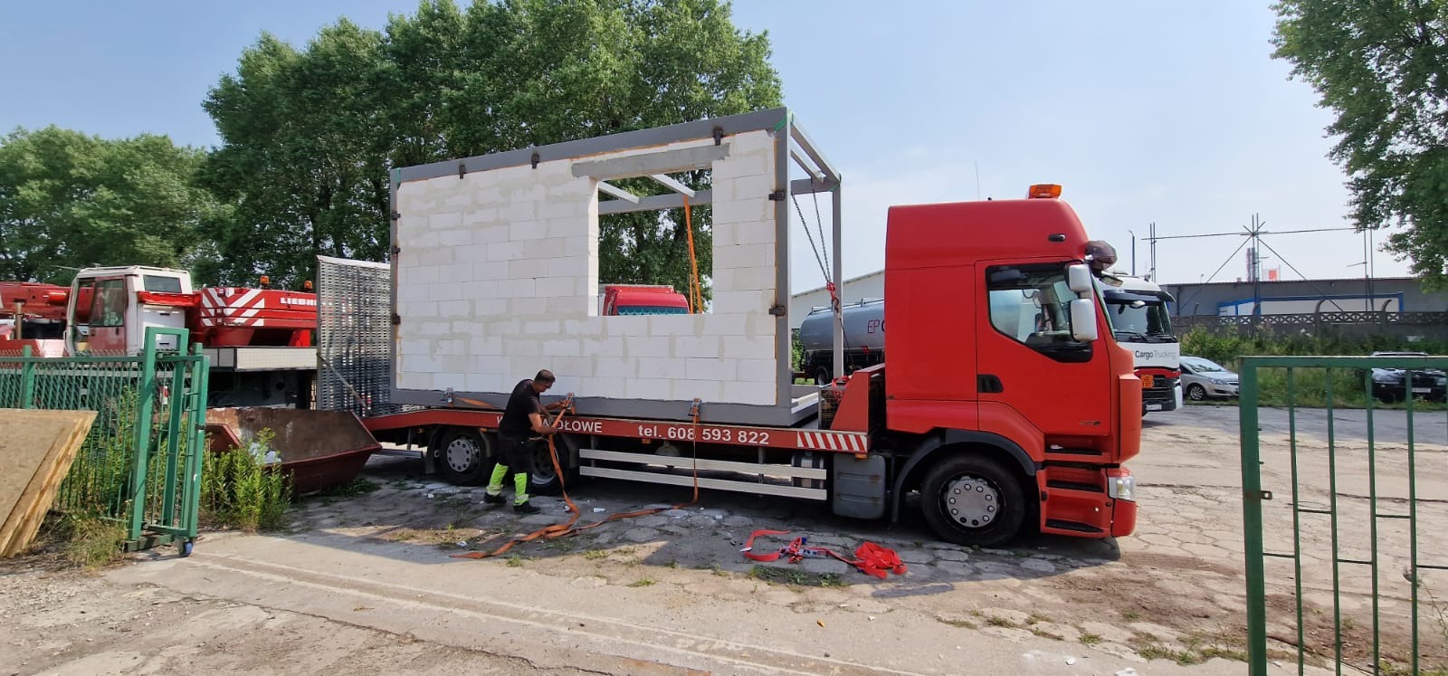 Transport modułu domu z bloczków betonowych na lawecie czerwonej ciężarówki, zabezpieczanego przez pracownika w odblaskowej kamizelce, z dźwigiem i innymi pojazdami w tle.