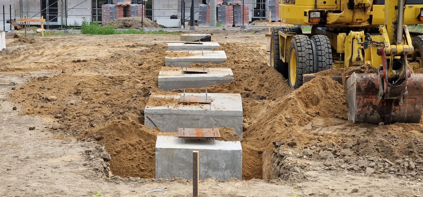 Przygotowanie fundamentów pod budowę domu: wykopane rowy z betonowymi stopami fundamentowymi, widoczna koparka Komatsu.