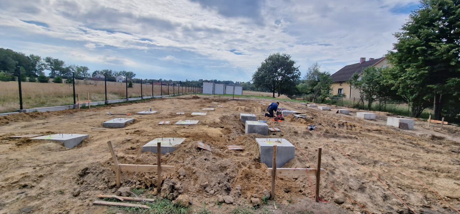 Przygotowanie terenu pod budowę domu w Katowicach: widoczne betonowe fundamenty z wystającymi prętami zbrojeniowymi, wykopane rowy, pracownik używający elektronarzędzia oraz ogrodzenie w tle.