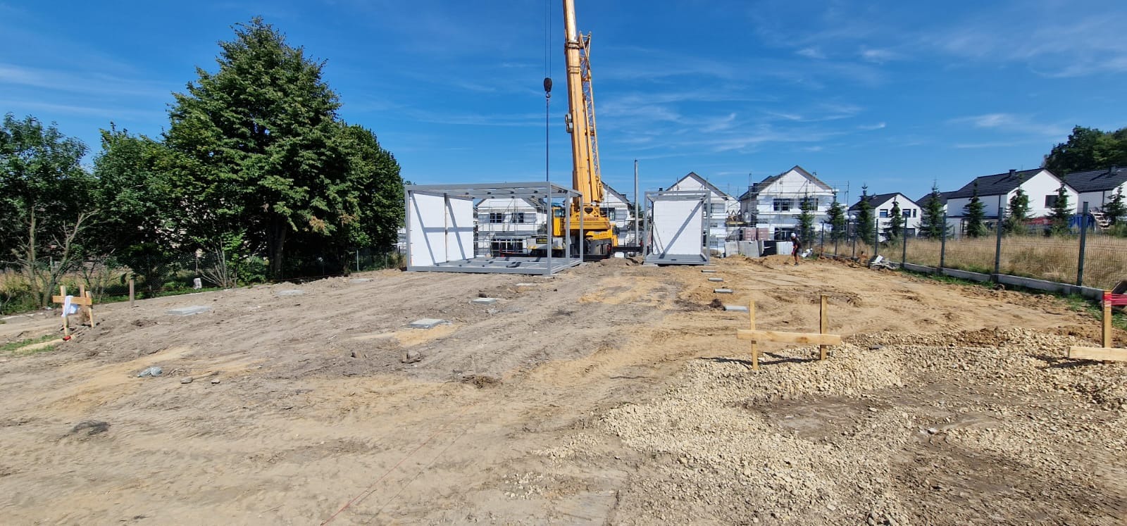 Montaż szkieletu modułowego domu na przygotowanym terenie budowy, widoczny dźwig oraz fundamenty, w tle zabudowa jednorodzinna.