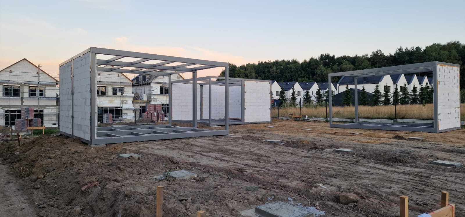 Wczesny etap budowy kilku domów modułowych z szarym szkieletem i białymi bloczkami na tle innych domów w budowie i drzew.