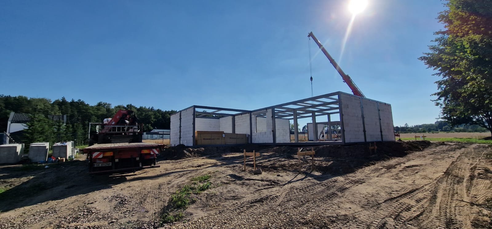 Wznoszenie szkieletu domu z prefabrykatów na fundamencie, widoczny żuraw i materiały budowlane na placu budowy w słoneczny dzień.