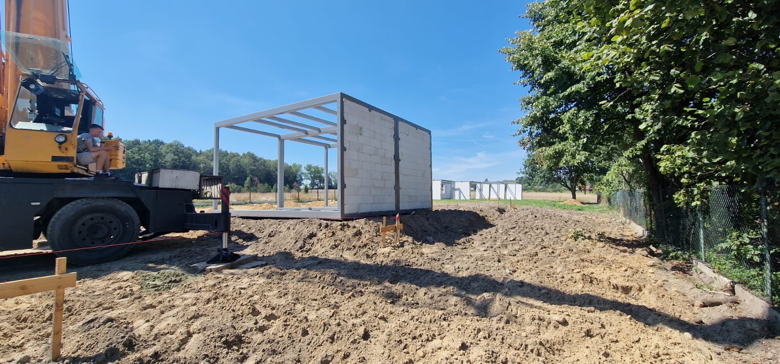 Montaż szkieletu domu z bloczków betonowych i stalowej konstrukcji nośnej przy użyciu dźwigu, widok na plac budowy w słoneczny dzień.