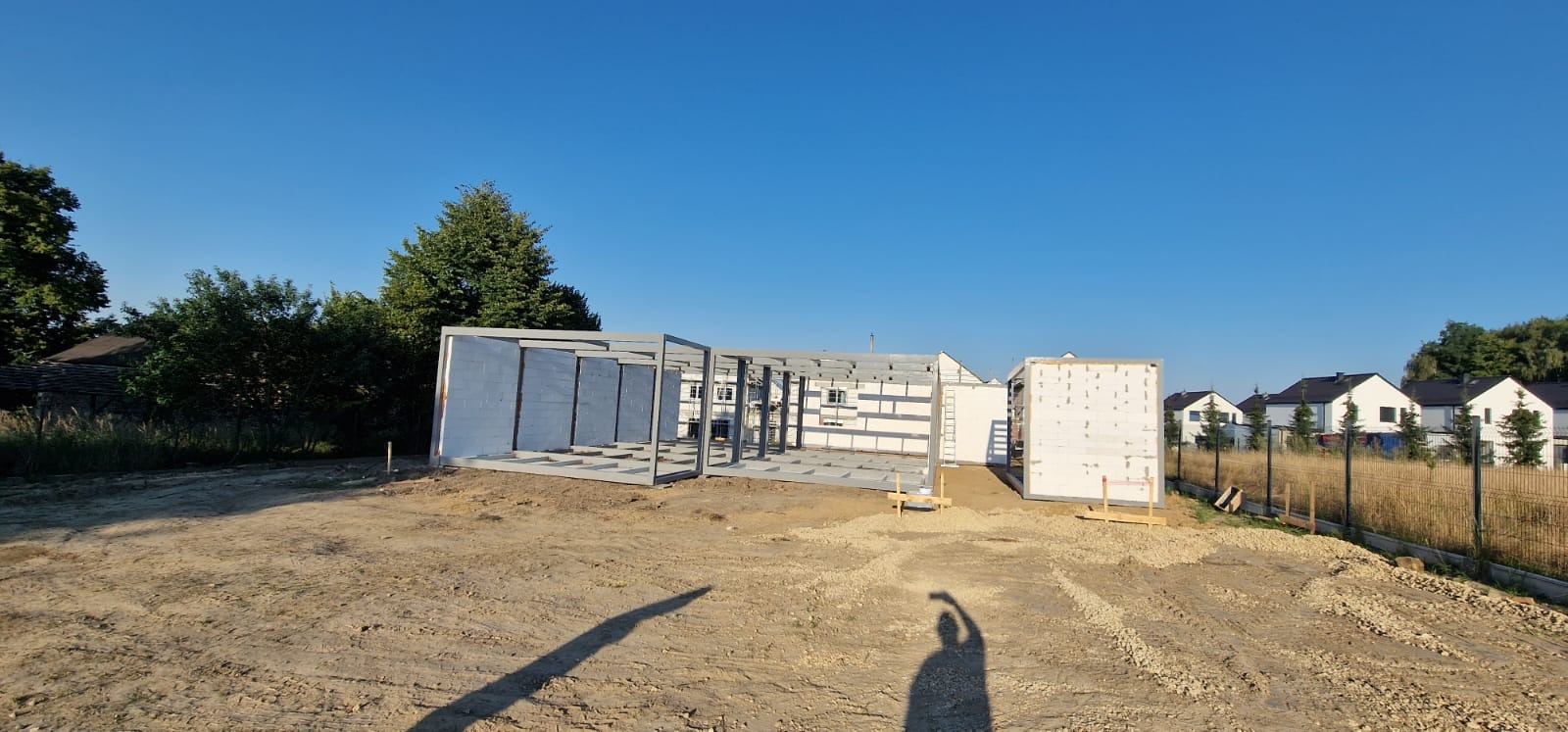 Wczesny etap budowy domu modułowego: widoczna stalowa konstrukcja szkieletowa i panele ścienne na przygotowanym placu budowy, w tle domy jednorodzinne.