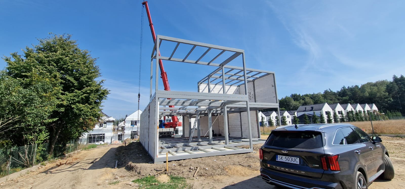 Dwukondygnacyjny dom modułowy w trakcie montażu, widoczny stalowy szkielet konstrukcyjny, żuraw budowlany i samochód Kia Sorento na pierwszym planie.
