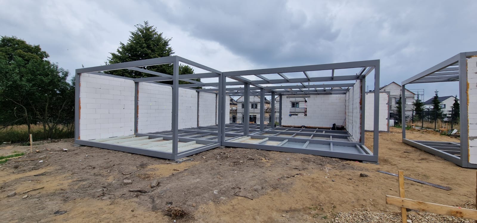 Konstrukcja modułowa w trakcie budowy: szkielet z belek i ścian z białych bloczków, widoczne fundamenty i otoczenie placu budowy w pochmurny dzień.