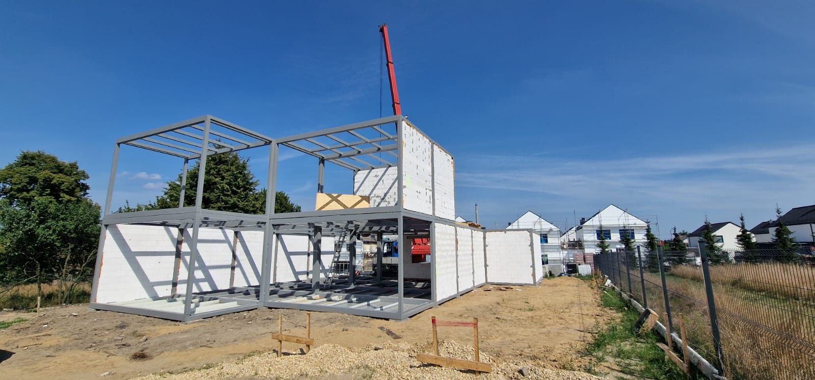 Wznoszenie dwukondygnacyjnego domu z modułów, widoczna stalowa konstrukcja szkieletowa i białe ściany z betonu komórkowego, w tle sąsiednie domy i żuraw budowlany.