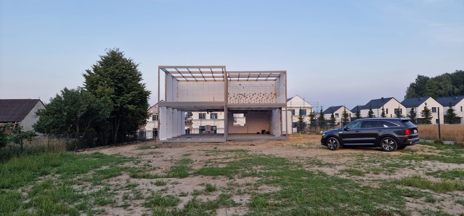 Surowy szkielet dwukondygnacyjnego domu z betonu komórkowego w trakcie budowy, z widocznym samochodem osobowym i okoliczną zabudową.