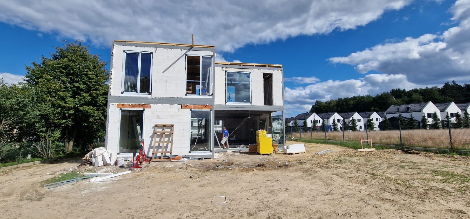 Dwukondygnacyjny dom w stanie surowym otwartym z białego bloczka, widoczne okna bez szyb, paleta z materiałami budowlanymi, pracownik wewnątrz budynku, w tle inne domy jednorodzinne.