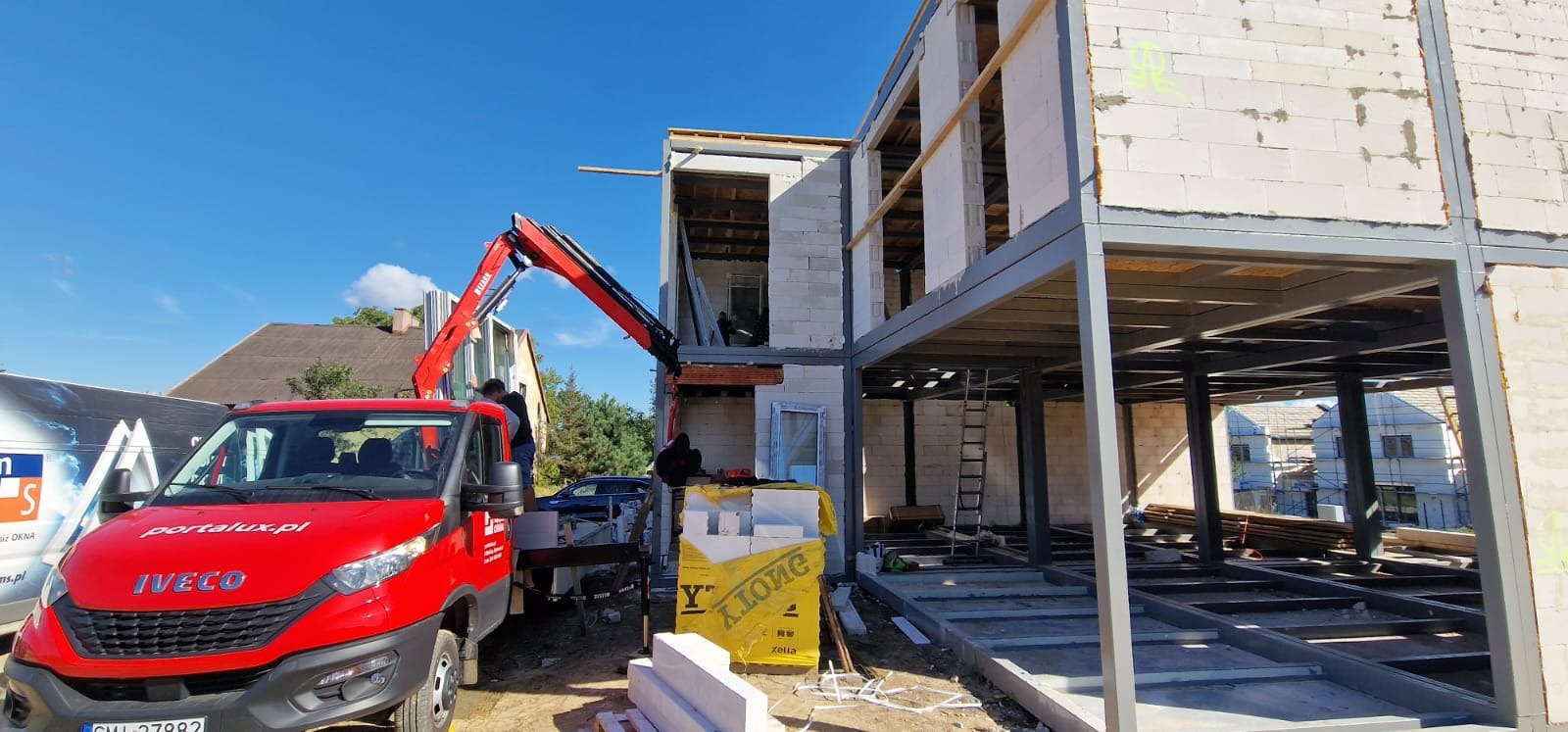 Budowa dwukondygnacyjnego domu z elementami stalowymi i bloczków Ytong, widoczny dźwig i samochód dostawczy z logo portalux.pl