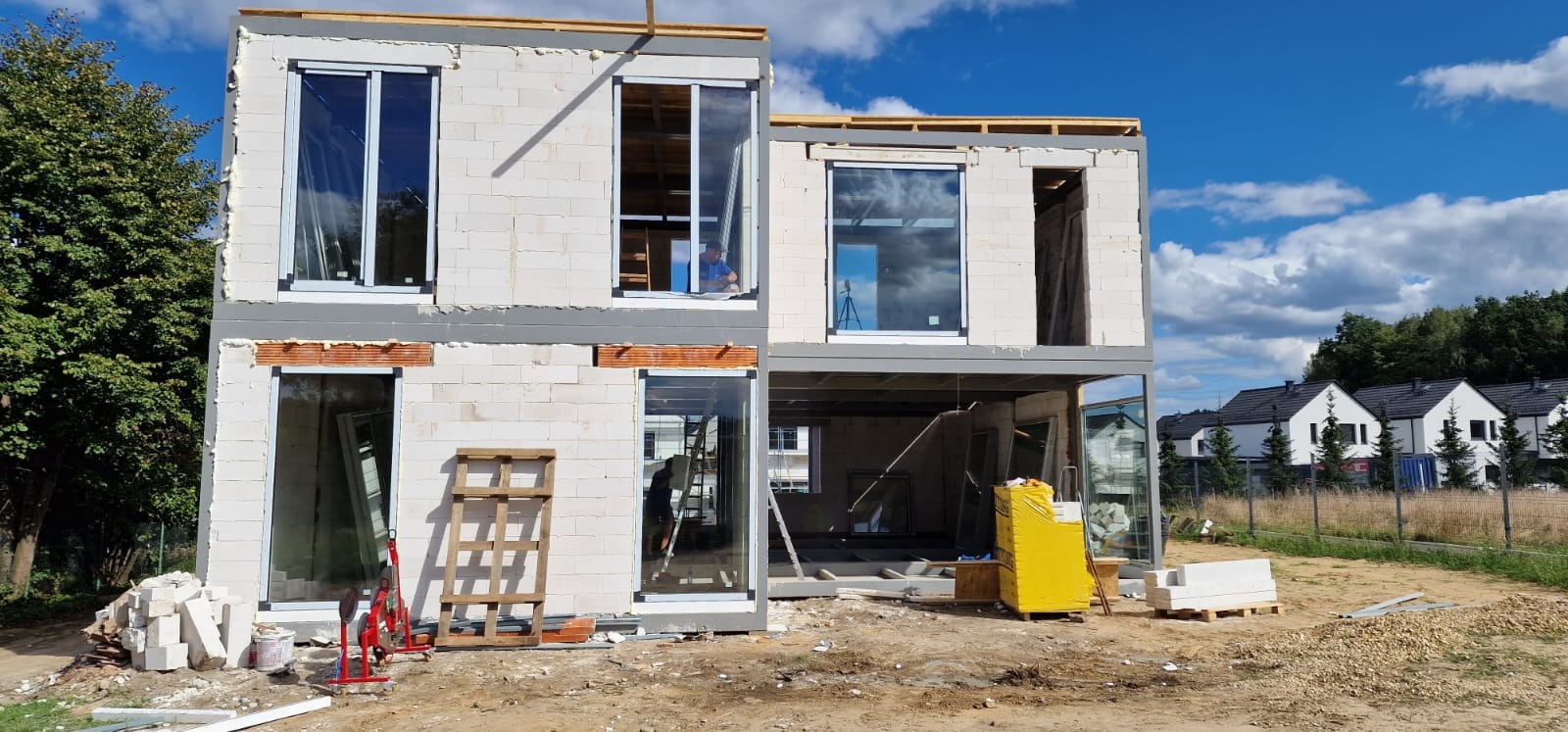 Dwukondygnacyjny dom w stanie surowym otwartym z białych bloczków, z zamontowanymi oknami, widoczne narzędzia i materiały budowlane, w tle domy jednorodzinne.