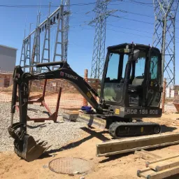 Czarna mini koparka Terex na placu budowy, widoczne elementy infrastruktury energetycznej w tle, ziemia i betonowe elementy na pierwszym planie.