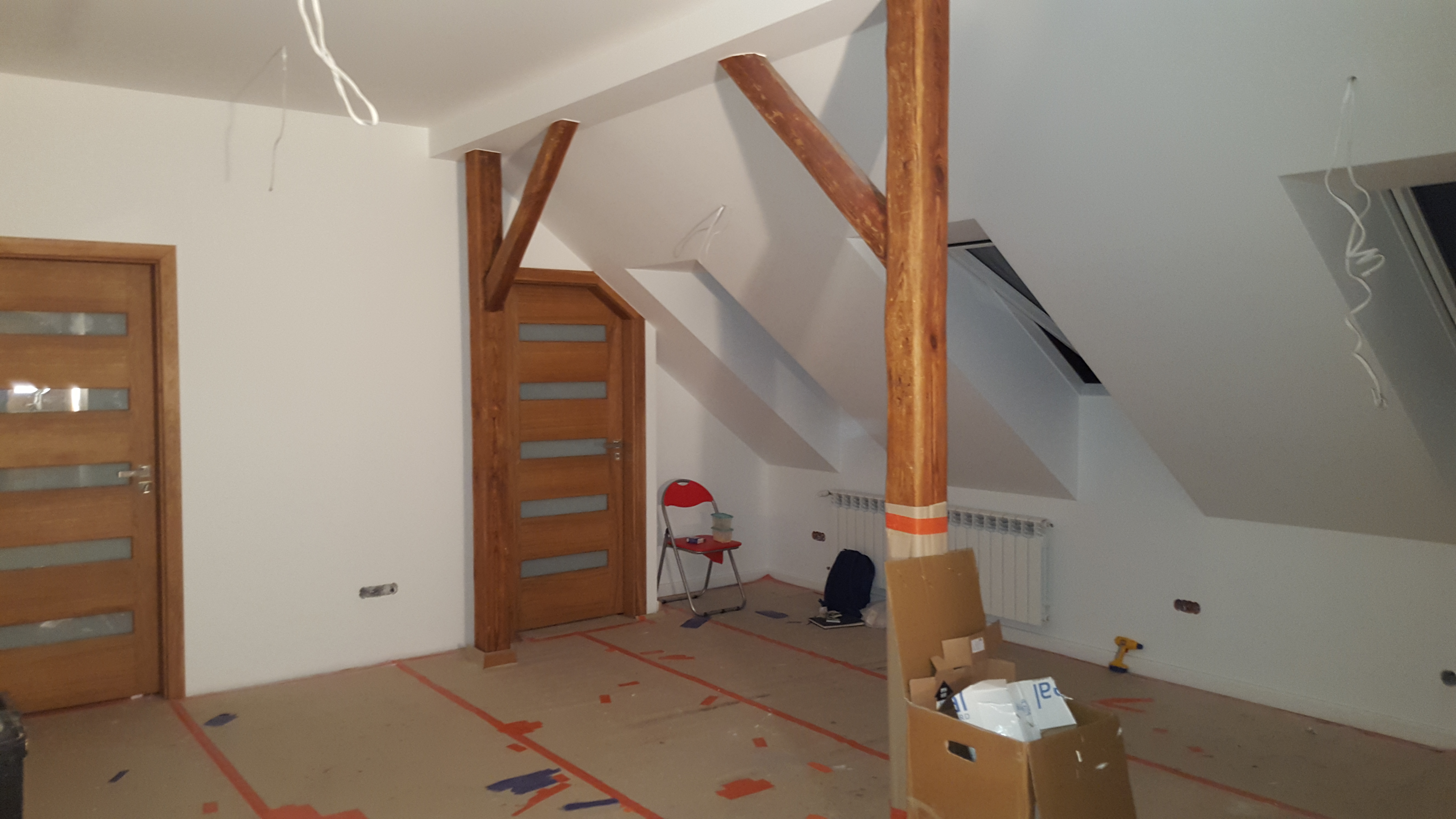 Pokój na poddaszu w trakcie remontu, zabezpieczona podłoga, drewniane elementy konstrukcyjne, widoczny grzejnik, okno dachowe i narzędzia.