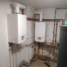 Dwa wiszące, białe piece centralnego ogrzewania marki Termet z białymi kominami, widoczne rury miedziane i szare, podłączone do instalacji, oraz szary zbiornik w tle. Na ścianach ślady wilgoci.