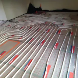 Rozłożone białe rurki ogrzewania podłogowego na folii z siatką, widoczne czerwone i niebieskie paski mocujące.